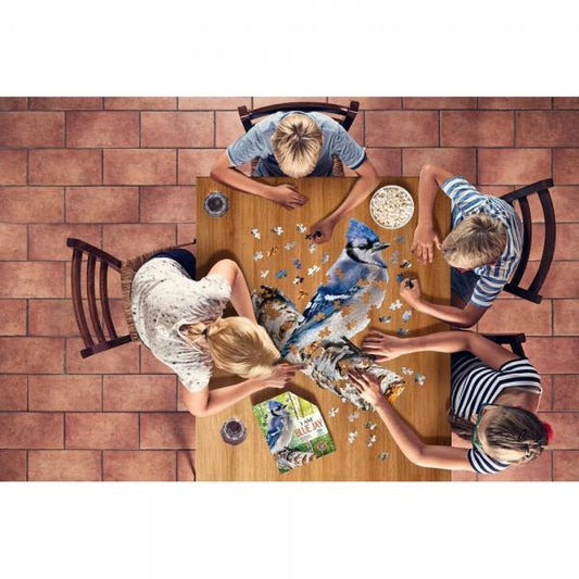 Overhead view of four children sitting around a wooden table playing with a jigsaw puzzle featuring a blue jay, with a puzzle box visible on the table.