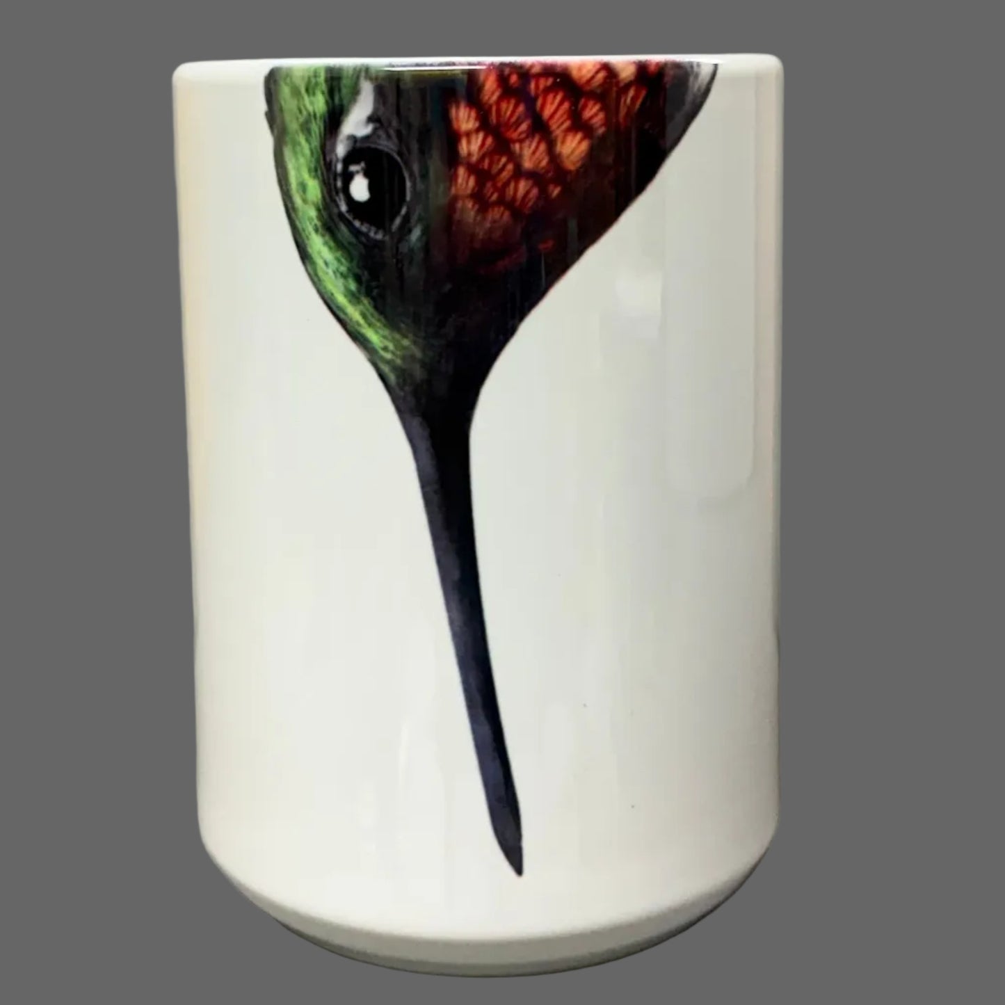 A white ceramic mug featuring a detailed illustration of a hummingbird with green, orange, and black feathers, set against a gray tablet case and wooden background.