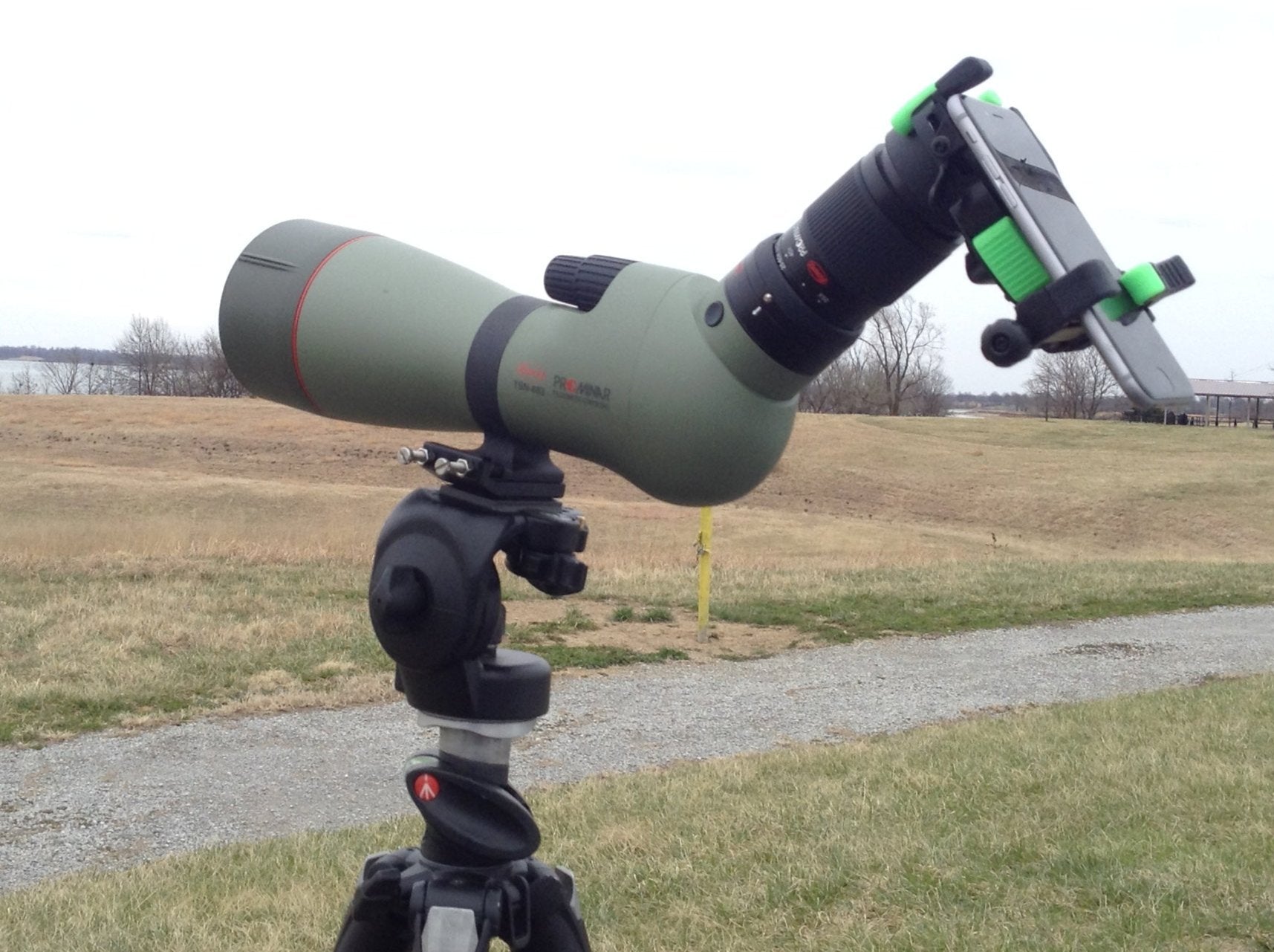 Black and green spotting scope with black tripod, phone adapter and smart phone.