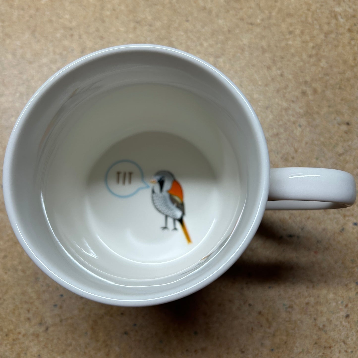 A white ceramic mug viewed from above, showing a small bird illustration inside with a speech bubble containing the text 'TTF'. The mug is placed on a textured brown surface.