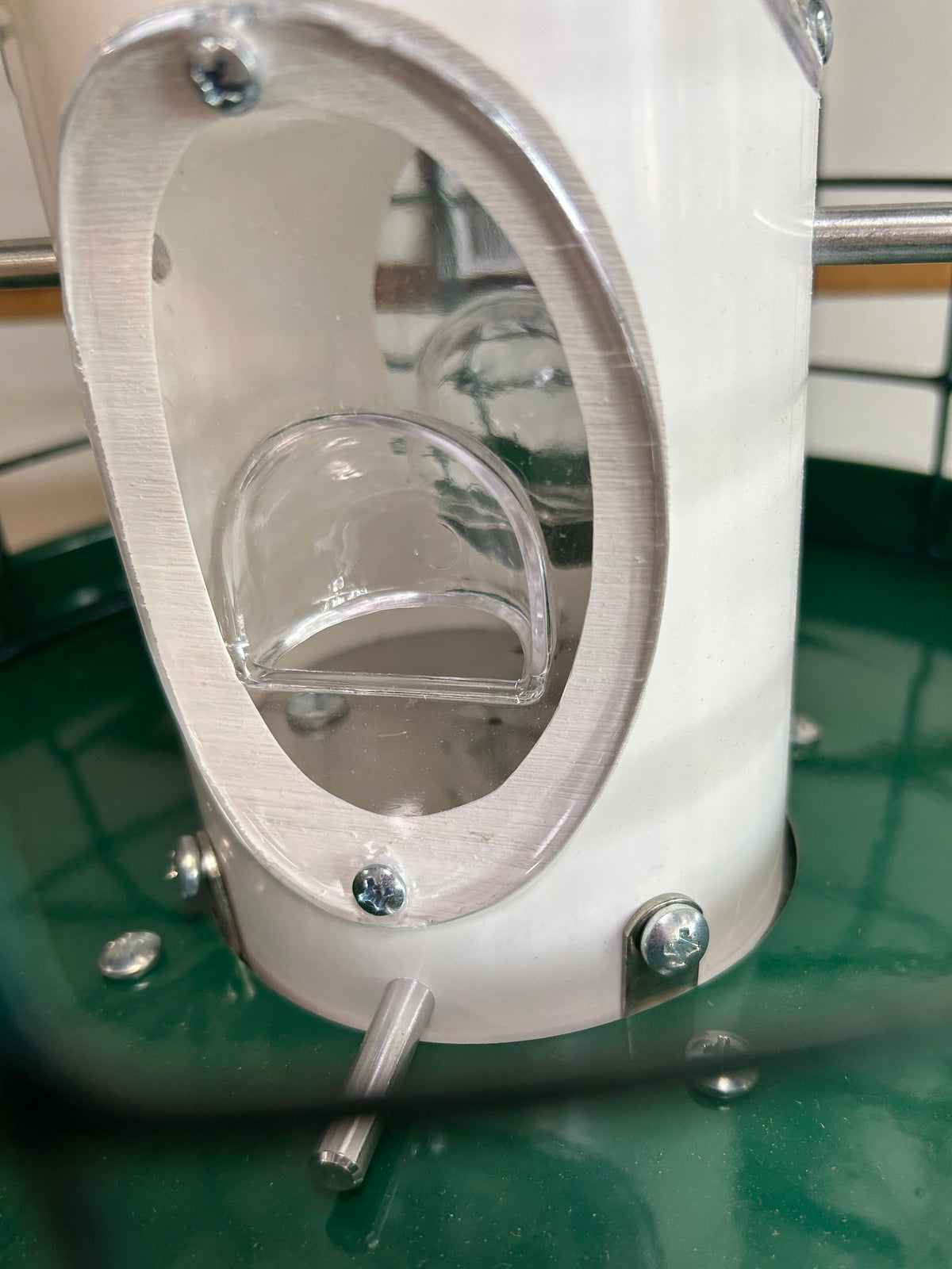 A close-up of a white cylindrical mechanical component with a clear plastic bowl or cup inside, mounted on a green surface with visible screws and metal rods.