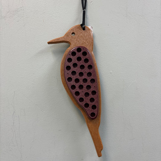 A wooden bird-shaped hanging ornament with a black hanging cord, featuring a natural wood body and a dark reddish-brown central panel covered in circular holes, shown against a light gray textured wall.