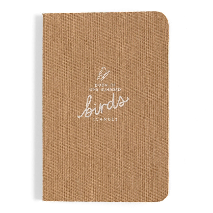 A tan linen-covered notebook with rounded corners, featuring white printed text and logo on the front reading 'Book of One Hundred Birds' and 'I CANOE!', shown on a clean white background.